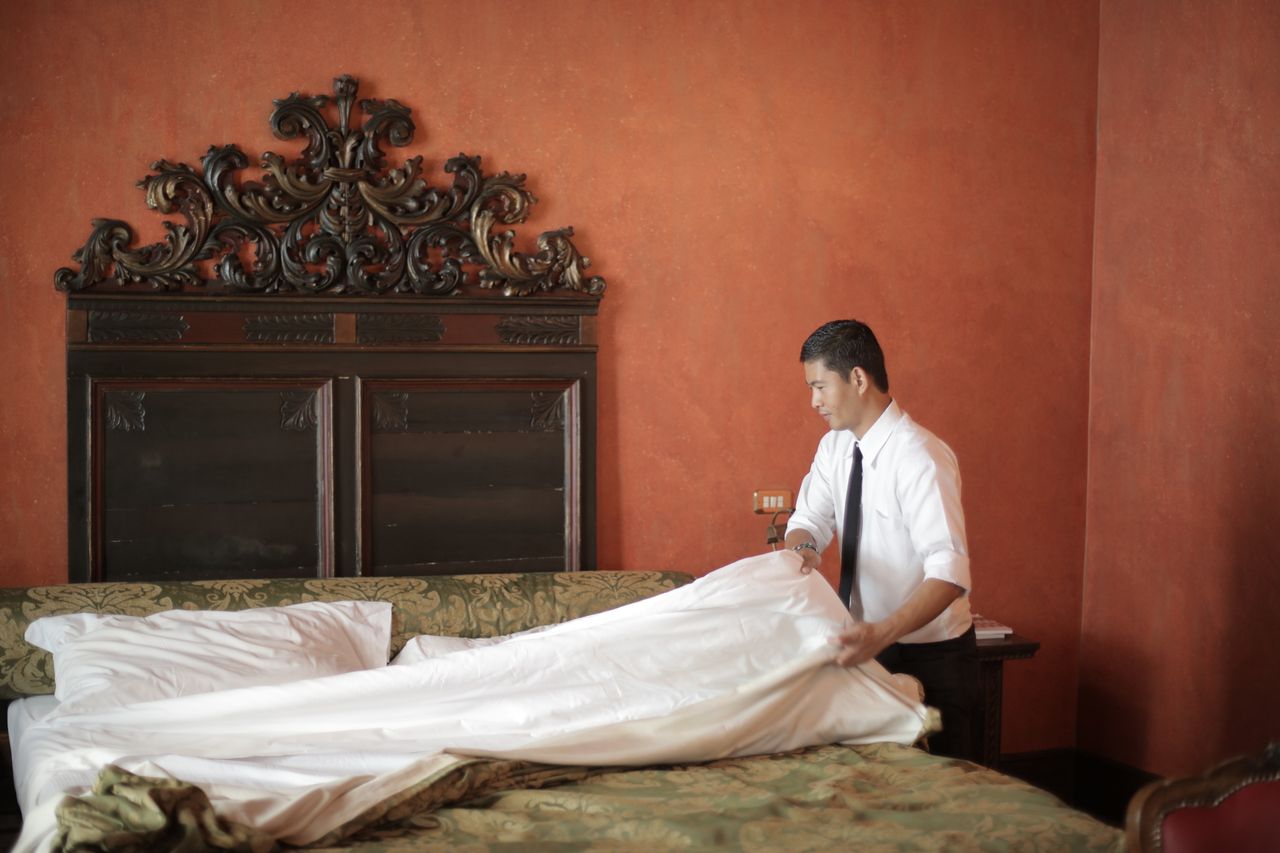 services-02 A hotel staff member in formal attire making a bed in a luxurious bedroom setting.